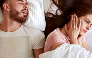 A man is lying in bed on his back, snoring loudly with his mouth open. Beside him, his wife is sitting up in bed, looking frustrated and tired, with her hand over her ear and a frown on her face.